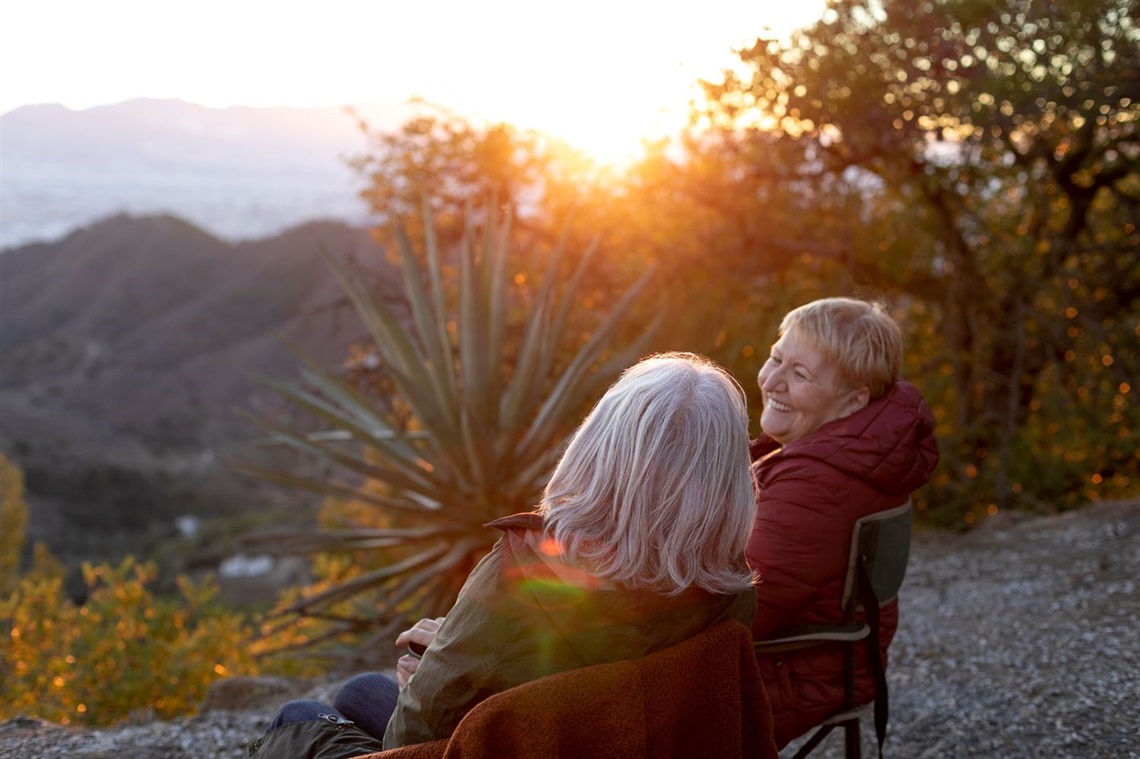 two elderly women sit on a bench on a mountain top during, with the sunset and a vista out in front of them. one woman has her head turned towards the other, smiling. 
