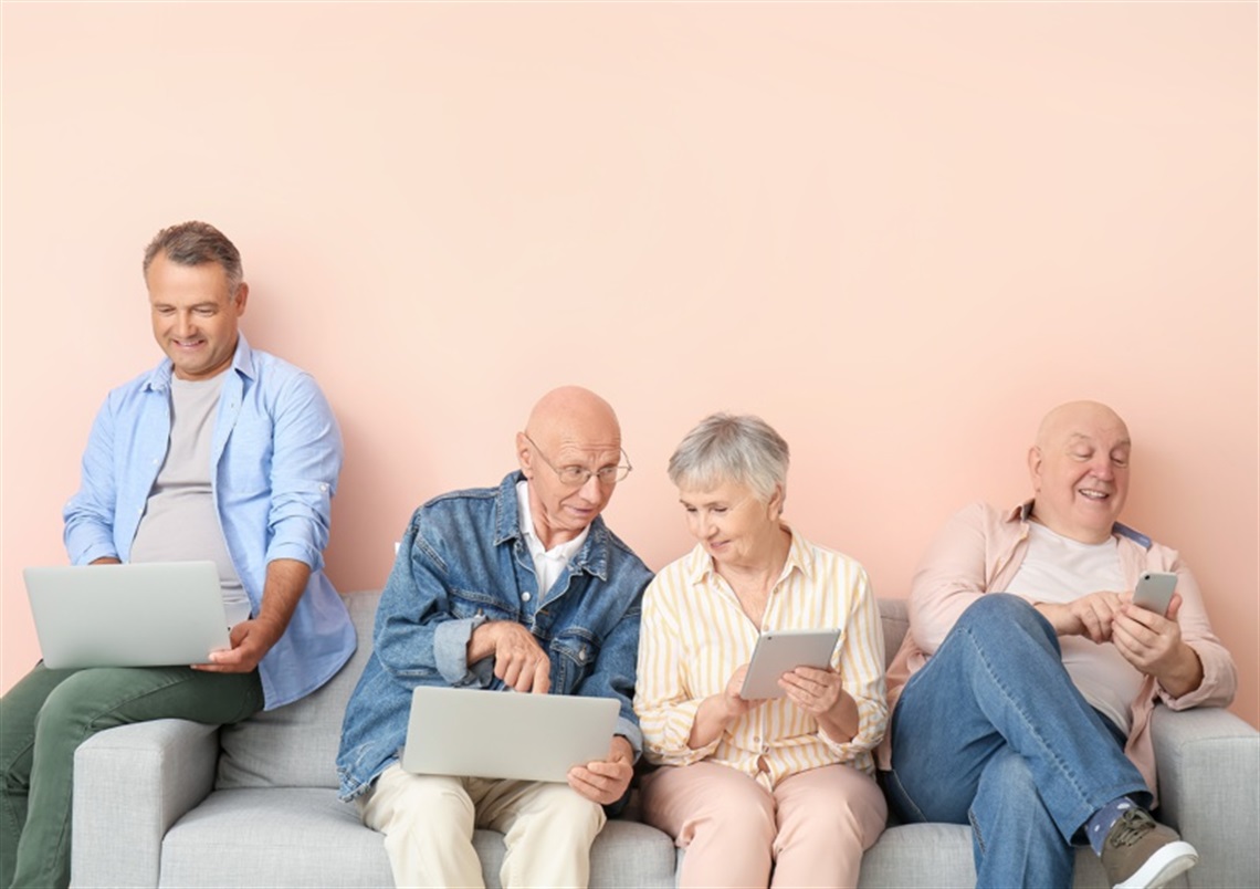 Four people using various Apple digital devices sitting on a grey couch with a pink wall behind